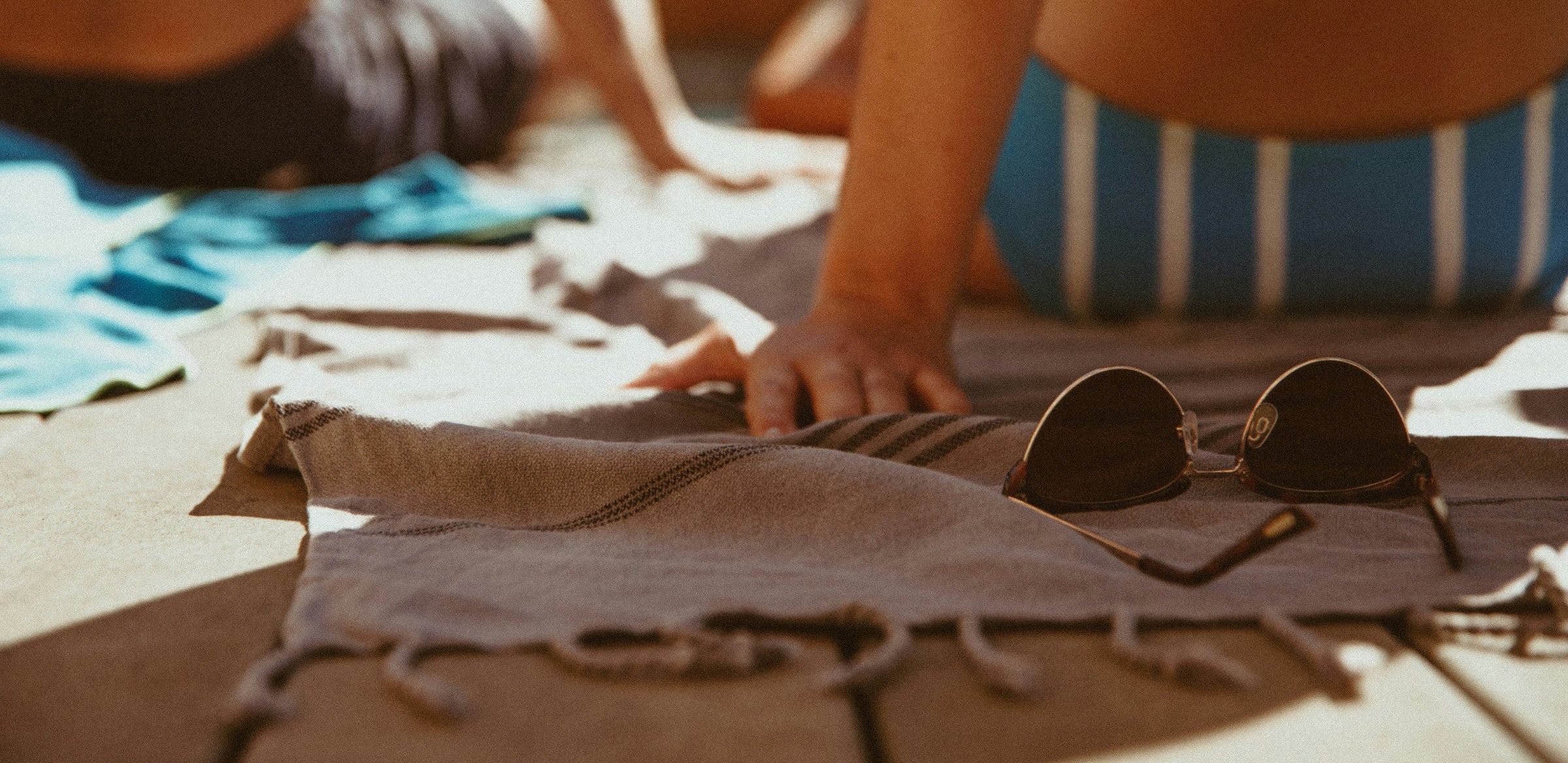 A close-up of a person’s arm resting on a towel next to a pair of sunglasses and Goodlather bar soap by a pool. Another person is blurred in the background, both wearing swimsuits and enjoying a sunny day outdoors.
