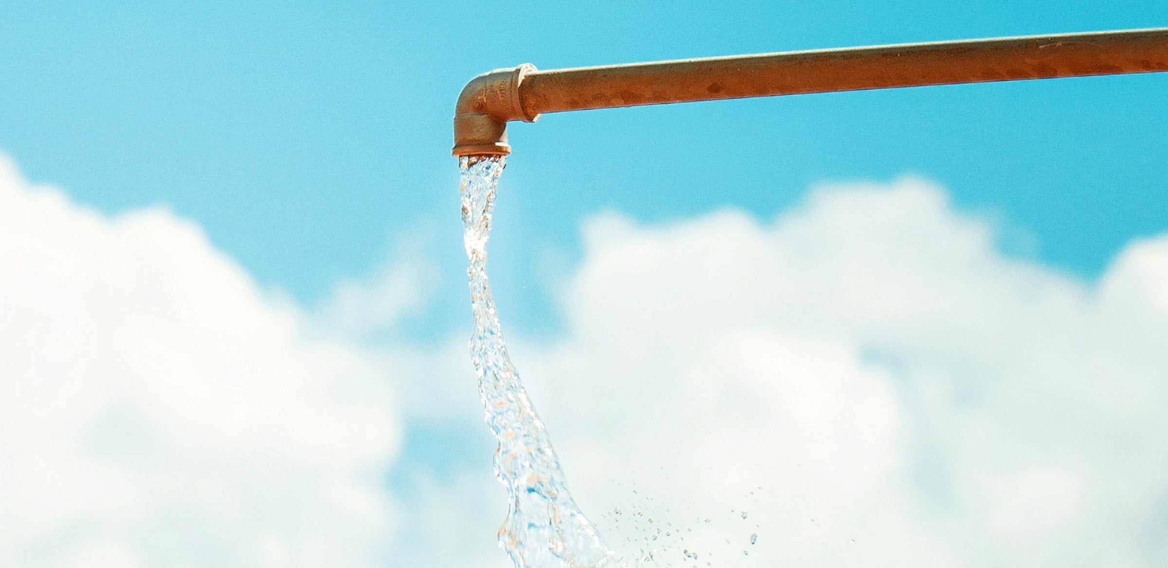 A close-up of water flowing from a rusty metal pipe—like an outdoor shower—against a bright blue sky with fluffy white clouds in the background.