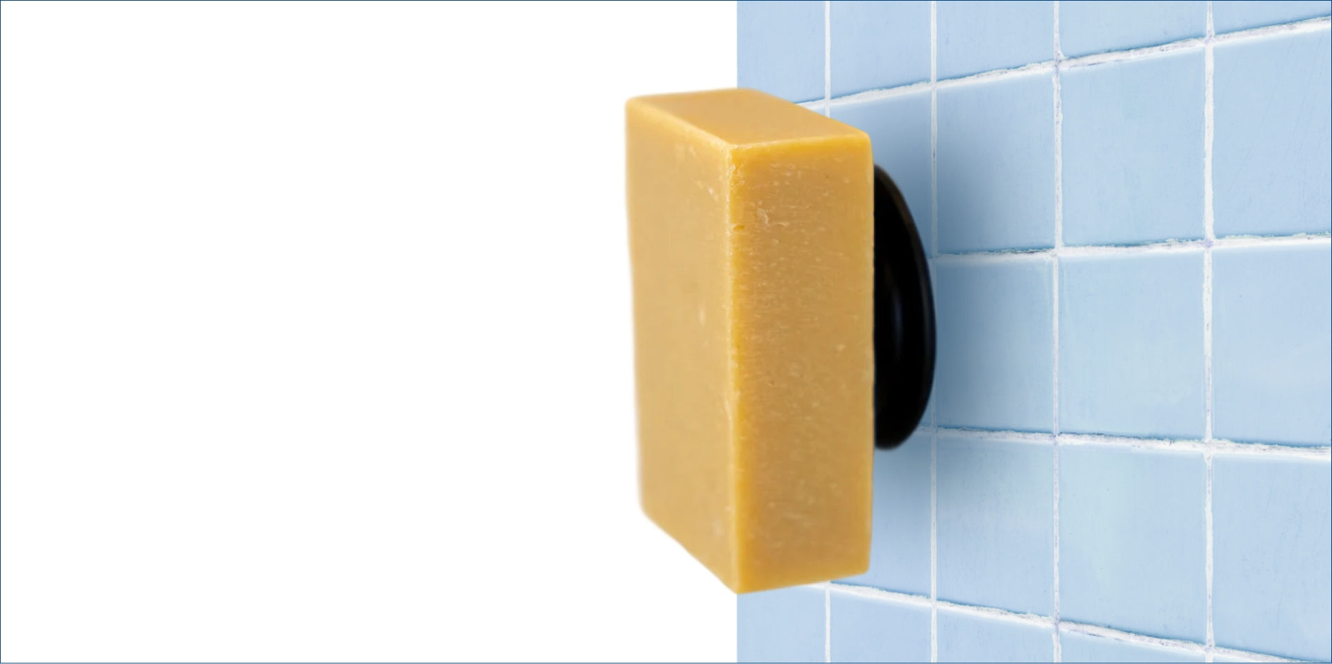 A rectangular bar of eco-friendly soap, made with natural ingredients, is magnetically attached to a round black holder on a blue tiled bathroom wall, appearing to float elegantly against the tiles.