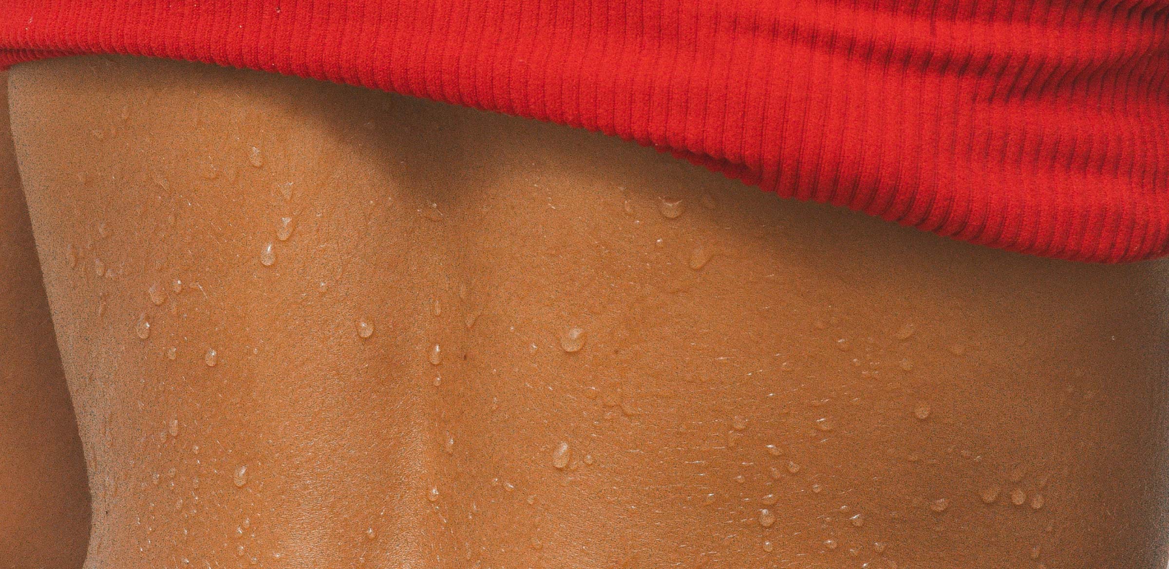 Close-up of a person's lower back with visible sweat droplets on their skin, partially covered by a red ribbed garment at the top—perfect for showing off that goodlather glow.